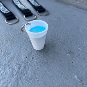Puede incluir: Un llavero de plástico blanco en forma de vaso con un líquido azul en su interior. El vaso tiene un anillo plateado.