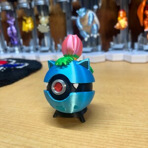 Puede incluir: Una figura de Pokemon Snorlax impresa en 3D de color azul y negro con una flor rosa en la parte superior. La figura est&aacute; de pie sobre una peque&ntilde;a base negra.
