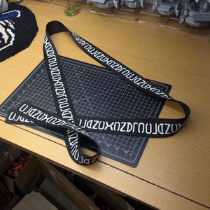 Może przedstawiać: Czarny pasek z białym napisem "JAZUXZNDJROUJZUX". Pasek leży na czarnej macie do cięcia na drewnianym stole. W tle kilka figurek w gablotach.