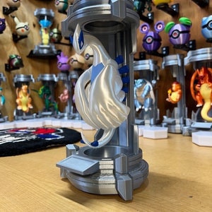 Puede incluir: Una figurita impresa en 3D de una criatura m&iacute;tica blanca con detalles azules, de pie sobre un pedestal gris con una superficie texturizada. La figurita est&aacute; en una pose con las alas extendidas.