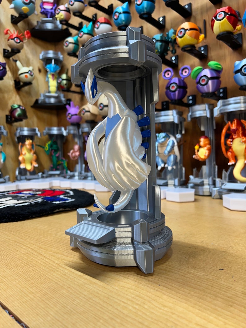 Puede incluir: Una figurita impresa en 3D de una criatura m&iacute;tica blanca con detalles azules, de pie sobre un pedestal gris con una superficie texturizada. La figurita est&aacute; en una pose con las alas extendidas.