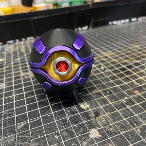 Puede incluir: Una esfera negra con un diseño dorado y morado. La esfera tiene un ojo rojo en el centro.