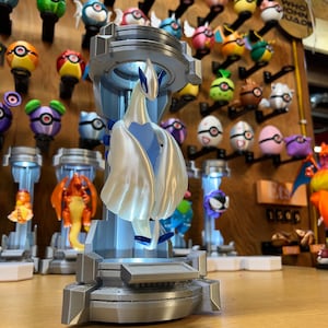 Puede incluir: Una figura impresa en 3D de un Pok&eacute;mon blanco parecido a un p&aacute;jaro con detalles azules, de pie sobre una plataforma gris. La figura est&aacute; iluminada desde abajo, creando un efecto brillante. La figura est&aacute; rodeada de otras figuras de Pok&eacute;mon sobre una superficie de madera.