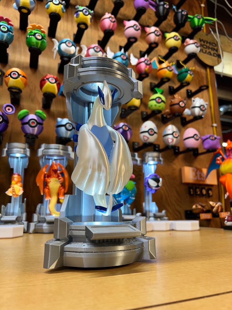 Puede incluir: Una figura impresa en 3D de un Pok&eacute;mon blanco parecido a un p&aacute;jaro con detalles azules, de pie sobre una plataforma gris. La figura est&aacute; iluminada desde abajo, creando un efecto brillante. La figura est&aacute; rodeada de otras figuras de Pok&eacute;mon sobre una superficie de madera.