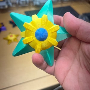 May include: A teal star-shaped object with a yellow center and a blue circle in the middle. The object is made of plastic and has a smooth surface.