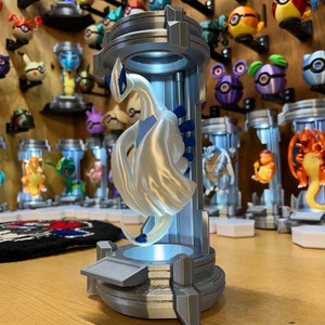 Puede incluir: Una figurita impresa en 3D de una criatura m&iacute;tica blanca con detalles azules, de pie sobre una plataforma gris con una luz en el interior. La figurita est&aacute; rodeada de otras figuritas impresas en 3D.