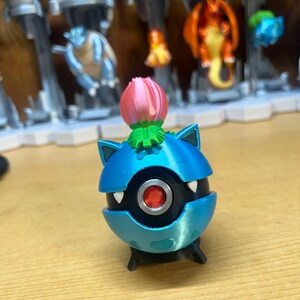 Puede incluir: Una figura de Pok&eacute;mon impresa en 3D de color azul y negro con una flor rosa en la parte superior. La figura tiene un ojo rojo y dientes blancos.