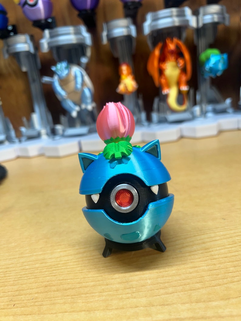 Puede incluir: Una figura de Pok&eacute;mon impresa en 3D de color azul y negro con una flor rosa en la parte superior. La figura tiene un ojo rojo y dientes blancos.
