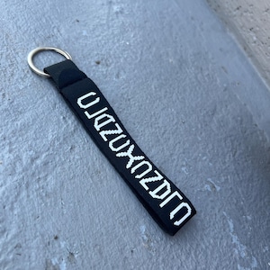 May include: Black keychain strap with white text and a silver metal ring. The text reads "OJZUXZDZO". The strap is made of a woven material and is likely used for keys or other small items.