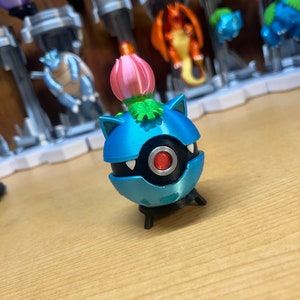Puede incluir: Una figura de Pok&eacute;mon azul y negro con una flor rosa en la parte superior. La figura est&aacute; de pie sobre una superficie de madera.