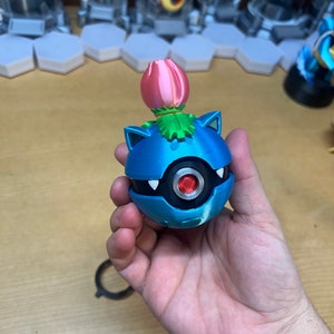 Puede incluir: Una figura de Pok&eacute;mon impresa en 3D de color azul y plateado con una flor rosa en la parte superior. La figura es una Pok&eacute; Ball con un ojo rojo y dientes blancos.
