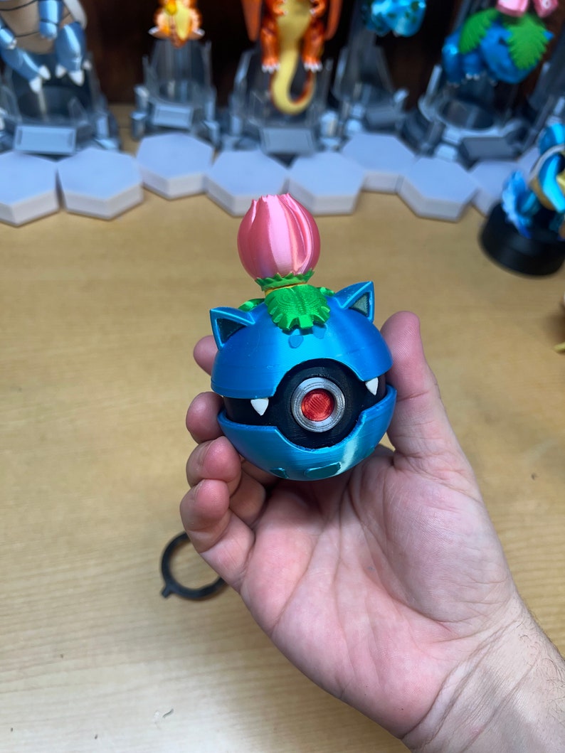 Puede incluir: Una figura de Pok&eacute;mon impresa en 3D de color azul y plateado con una flor rosa en la parte superior. La figura es una Pok&eacute; Ball con un ojo rojo y dientes blancos.