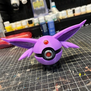 May include: A purple and black 3D printed figurine of a Pokemon character with large ears. The character has a red eye and a black ring around its eye.
