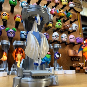 Puede incluir: Una figura impresa en 3D de un personaje Pokemon blanco y azul, de pie sobre una plataforma gris. La figura est&aacute; posada con las alas extendidas. El fondo presenta una pared con varias figuras de Pokemon.