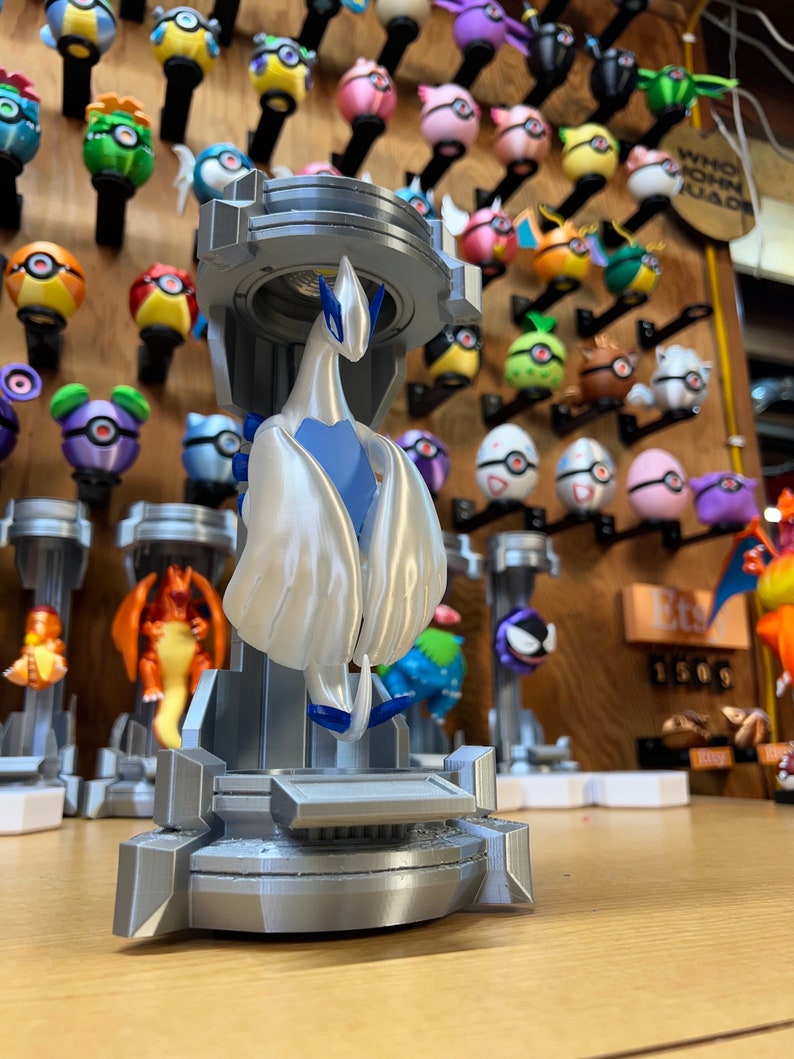 Puede incluir: Una figura impresa en 3D de un personaje Pokemon blanco y azul, de pie sobre una plataforma gris. La figura est&aacute; posada con las alas extendidas. El fondo presenta una pared con varias figuras de Pokemon.