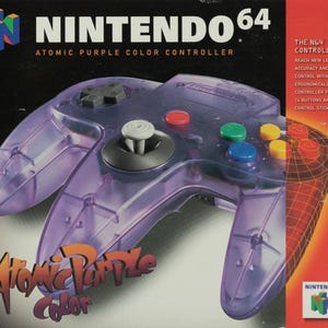 May include: Atomic Purple Nintendo 64 controller. The transparent purple controller has colored buttons and an analog stick. The box features the Nintendo 64 logo and text that reads "Atomic Purple Color Controller."