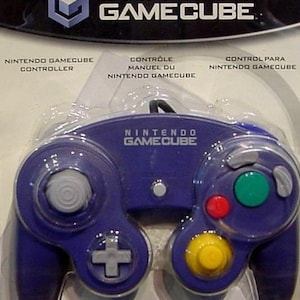 Może przedstawiać: Fioletowy kontroler Nintendo GameCube z białym i szarym analogowym joystickiem, żółtym przyciskiem, zielonym przyciskiem i czerwonym przyciskiem. Kontroler jest zapakowany w przezroczystą plastikową skorupę.