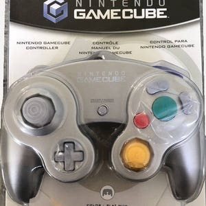 Może przedstawiać: Srebrny kontroler Nintendo GameCube w oryginalnym opakowaniu. Kontroler posiada szary pad kierunkowy, żółty drążek C, zielony przycisk A, czerwony przycisk B oraz szary przycisk Start/Pause. Na opakowaniu widnieje logo Nintendo GameCube.