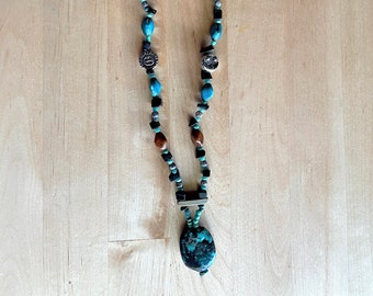 May include: A long necklace with turquoise, black, brown, and silver beads. The necklace has a large, dark turquoise pendant at the bottom and two silver charms. The beads are strung on a silver chain.