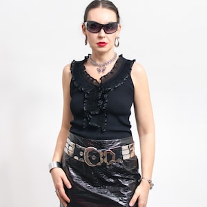 May include: A woman wearing a black sleeveless top with a ruffled neckline, a silver necklace, and a black shiny mini skirt. She is also wearing sunglasses, silver earrings, and a silver belt with a large buckle.