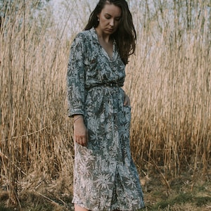 Op de afbeelding: Een jurk met lange mouwen en knopen, met een botanische print in grijstinten en wit. De jurk is in de taille getailleerd met een dunne, donkere riem. Het model draagt beige ballerina's. De jurk is knielang.