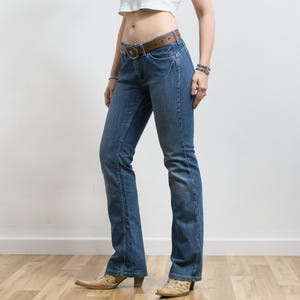 Peut inclure: Jean bleu avec une ceinture en cuir marron et une boucle d&eacute;corative. Le jean a une conception classique &agrave; cinq poches et une l&eacute;g&egrave;re jambe &eacute;vas&eacute;e. La personne porte des bottes de cowboy beiges &agrave; bout pointu.