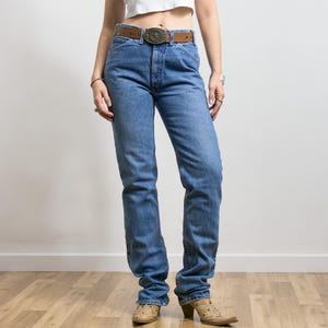 May include: Light blue denim jeans with a brown leather belt and a decorative buckle. The jeans have a straight-leg fit. The model is wearing cowboy boots.