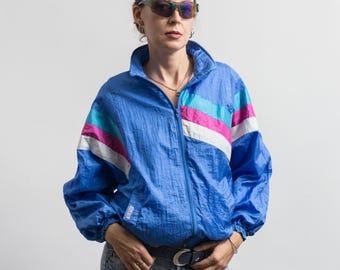 Peut inclure: Un coupe-vent bleu vif doté d'un col montant, d'une fermeture éclair intégrale sur le devant et de manches longues à poignets élastiques. La veste est rehaussée de bandes horizontales bleu sarcelle, magenta et blanches sur la poitrine.