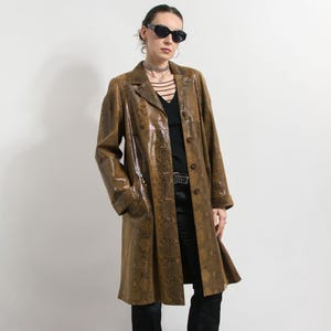 Könnte beinhalten: Ein langer, brauner Mantel mit Schlangenmuster, mit Reverskragen und Knopfverschluss. Der Mantel wird über einem schwarzen Oberteil und einer Hose getragen. Die Person trägt eine schwarze Sonnenbrille und eine silberne Halskette.