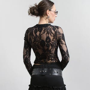 Puede incluir: Top negro de manga larga de encaje con cuello alto. El top se combina con una falda negra y un cinturón ancho negro con tachuelas plateadas. El conjunto es de estilo gótico o alternativo.