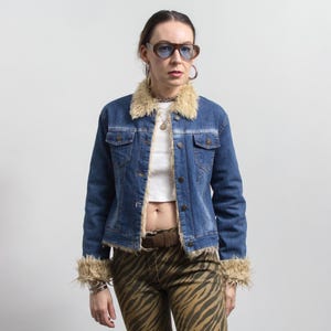 Peut inclure: Veste en jean bleue avec col et poignets en fausse fourrure beige. La veste a une fermeture boutonn&eacute;e et deux poches poitrine. La personne porte un pantalon z&eacute;br&eacute; marron et beige et des lunettes de soleil marron.