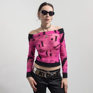 Pu&ograve; includere: Un top rosa acceso, con spalle scoperte, con finiture nere e maniche lunghe. Il top presenta un motivo di testo nero ripetuto. La modella indossa occhiali da sole neri, una collana d'argento e una cintura d'argento con pantaloni neri.
