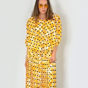 May include: A yellow and white polka dot dress with a pleated skirt. The dress has a knotted detail at the waist. The model is wearing black platform heels and yellow sunglasses.