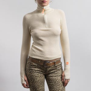 May include: A cream-colored ribbed knit sweater featuring a high collar with a half-zip closure, long sleeves with thumb holes, and a small orange logo patch on the left cuff. The sweater is paired with brown leopard print pants and a brown leather belt.