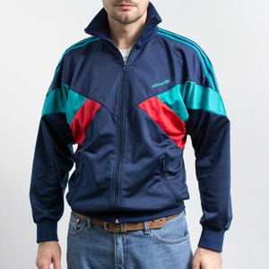 Puede incluir: Una chaqueta deportiva azul marino con cuello alto, cremallera frontal completa y mangas largas con tres rayas. El pecho presenta un patrón de bloques de color turquesa y rojo con el logotipo de Adidas bordado en turquesa en el lado izquierdo.
