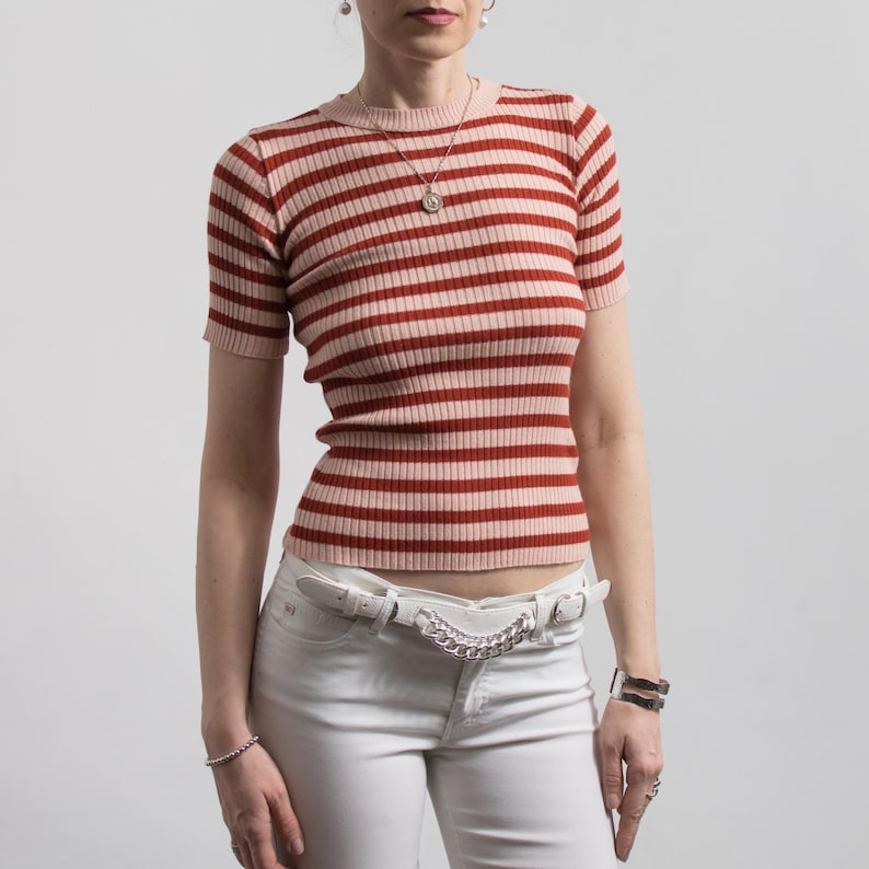 May include: A short-sleeved, ribbed knit top with horizontal stripes in shades of red and pink. The top is paired with white jeans and a white belt with a silver chain detail. The model is wearing jewellery.