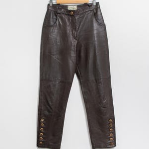 May include: Dark brown leather pants hanging on a wooden hanger. The pants have a high waist, pockets, and decorative gold buttons along the outer ankles. The leather appears soft and has a slight sheen.
