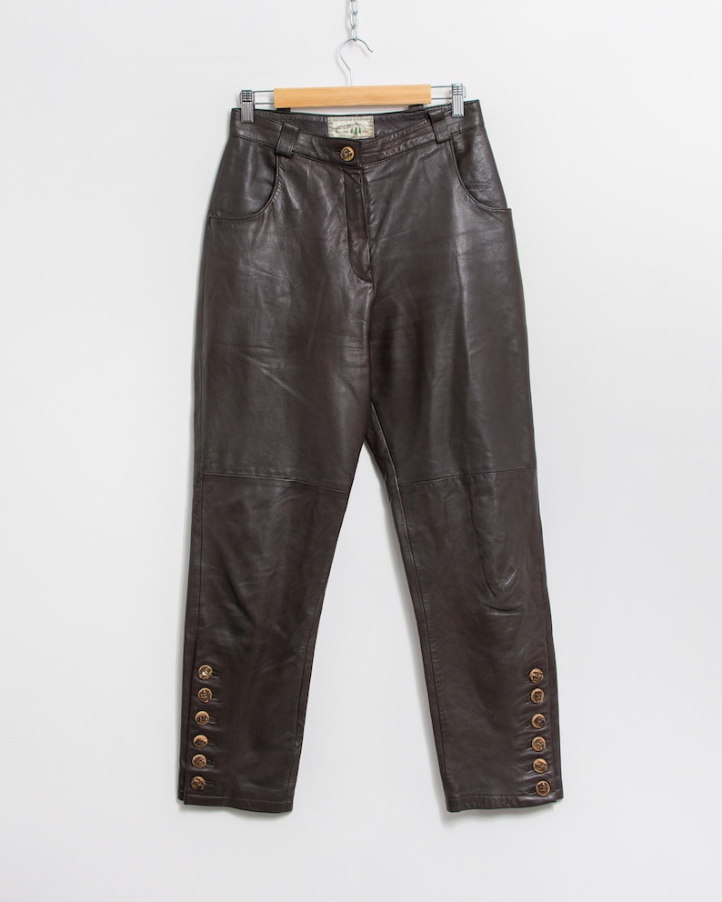 May include: Dark brown leather pants hanging on a wooden hanger. The pants have a high waist, pockets, and decorative gold buttons along the outer ankles. The leather appears soft and has a slight sheen.