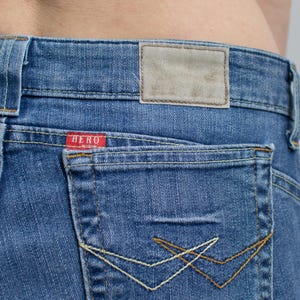 Peut inclure: Gros plan sur un jean en denim bleu avec une poche arri&egrave;re et une petite &eacute;tiquette rouge avec le mot "HERO". Le jean est d&eacute;lav&eacute; et poss&egrave;de une &eacute;tiquette en cuir &agrave; la taille. La poche a des coutures d&eacute;coratives de couleur contrastante.