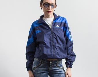Peut inclure: Un coupe-vent zippé bleu marine et bleu vif avec le logo Adidas blanc sur la poitrine, des manches longues à poignets élastiques, un col montant et deux poches avant zippées, porté sur un haut blanc à côtes.