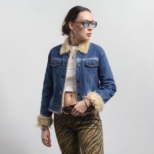 Peut inclure: Veste en jean bleue avec un col et des poignets en fausse fourrure marron clair. La veste a des poches boutonn&eacute;es et une fermeture &agrave; boutons. La personne porte des lunettes de soleil teint&eacute;es marron et un pantalon &agrave; imprim&eacute; z&eacute;br&eacute;.