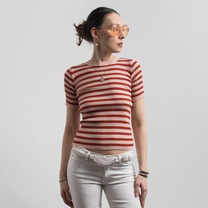 May include: A short-sleeved, ribbed knit top with horizontal red and pale pink stripes. The top is worn with white jeans and a white belt with a silver chain detail. The person is wearing orange-tinted sunglasses and a necklace.