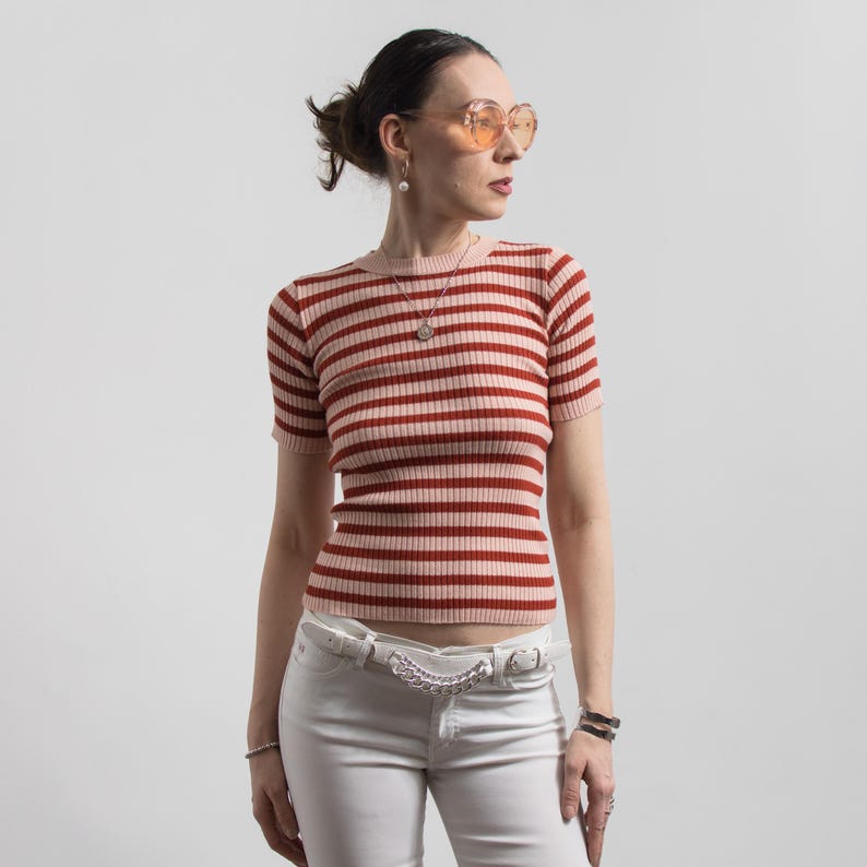 May include: A short-sleeved, ribbed knit top with horizontal red and pale pink stripes. The top is worn with white jeans and a white belt with a silver chain detail. The person is wearing orange-tinted sunglasses and a necklace.