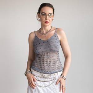 May include: A grey crocheted camisole top with a V-neck and spaghetti straps. The top features a dotted pattern and is worn with a white skirt and silver accessories, including a chain belt and bracelets.