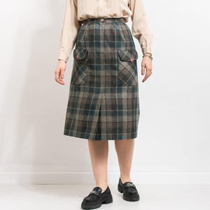Op de afbeelding: Een knielange geruite rok in tinten grijs, blauwgroen en bruin. De rok heeft twee voorzakken met knoopsluitingen en een voorste plooi. De stof heeft een getextureerd uiterlijk, wat wijst op een wolmix. De rok wordt gecombineerd met zwarte loafers.