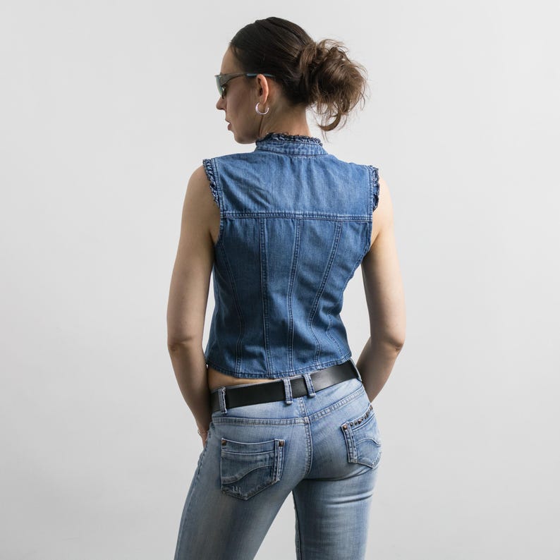 May include: A sleeveless blue denim vest featuring a ruffled collar and vertical seam detailing, paired with light blue denim jeans that have a black leather belt with a silver buckle. The vest has a fitted silhouette and a button-up front closure.