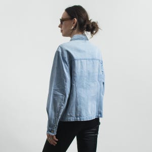May include: Light blue denim jacket with button closures and a classic collar. The jacket has a faded wash and a tailored fit. The sleeves have buttoned cuffs. The model is wearing black pants and sunglasses.