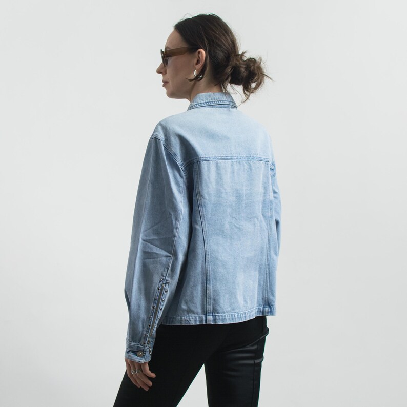 May include: Light blue denim jacket with button closures and a classic collar. The jacket has a faded wash and a tailored fit. The sleeves have buttoned cuffs. The model is wearing black pants and sunglasses.