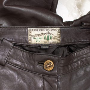 May include: Close-up of brown leather pants. The pants feature a button closure with a decorative gold button. A label reads "Natural Life" with a mountain and tree design. The size tag reads "40".