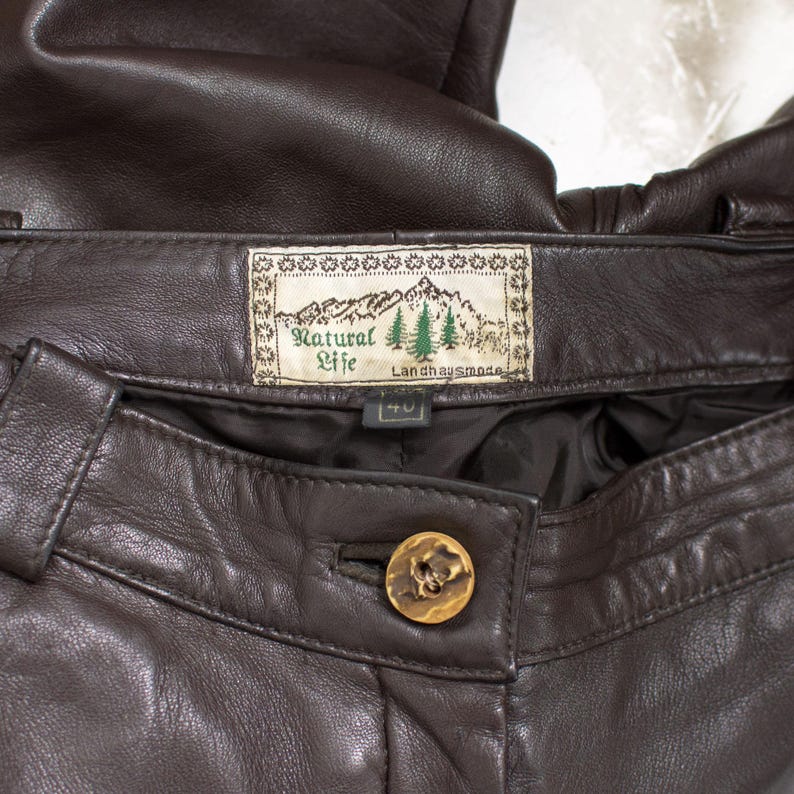 May include: Close-up of brown leather pants. The pants feature a button closure with a decorative gold button. A label reads "Natural Life" with a mountain and tree design. The size tag reads "40".